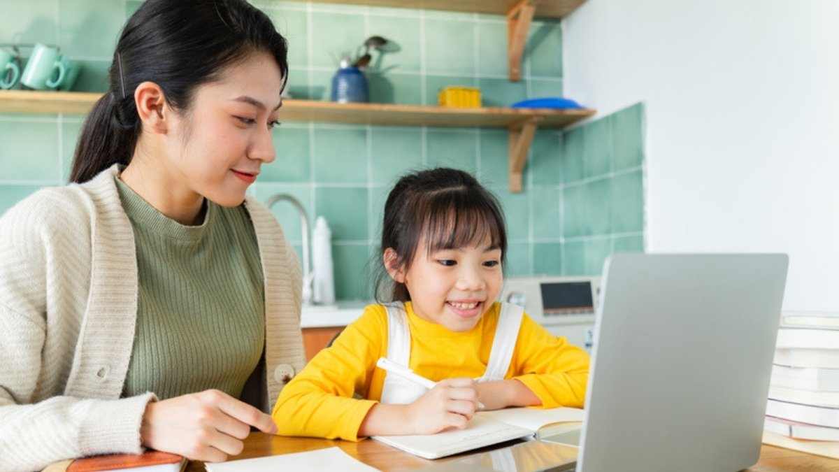A mother sits beside her smiling daughter as she writes in a workbook at the kitchen table